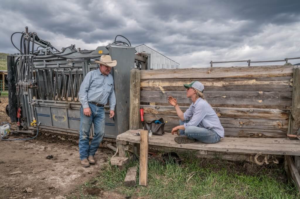 An older man is leaning against a cattle chute as a young woman who is sitting talks to him