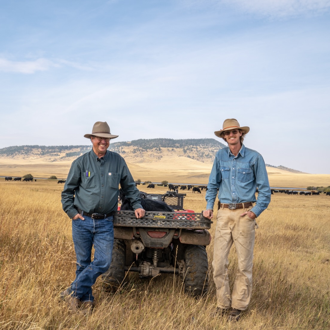 Two men leaning on an ATV, with a large herd of cows in a field behind them